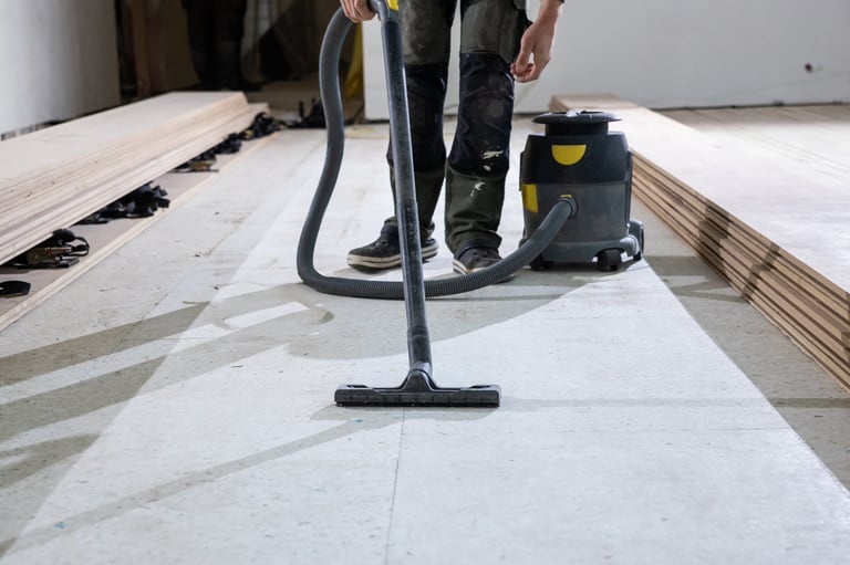 A worker uses a vacuum cleaner to clean a floor before installing wooden floorboards. Stacks of planks are visible, indicating a renovation project.